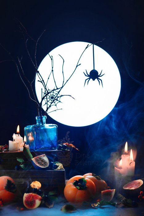 A scary still life featuring the silhouette of a spider, a full moon, candles and other spooky props