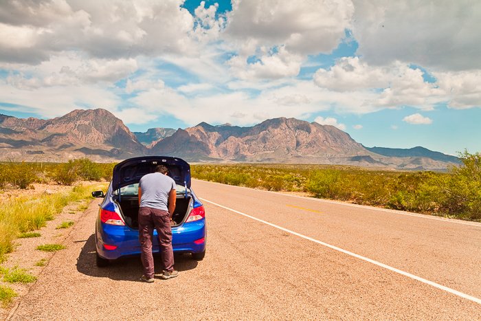 Man looking into the trunk of his blue car, stopped at the side of a pale brown road leading to the mountains in the distance