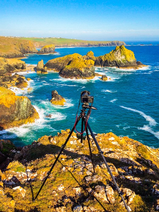 DSLR set up on a tripod at the top of brown rocks overlooking a rocky shore to a deep blue sea