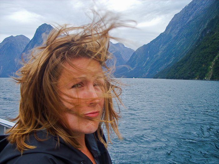 A woman taking a selfie in front of a blue lake, sky and mountains, the wind blowing her short blonde hair in her face