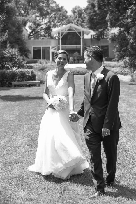 black and white photo of newly weds bride and groom holding hands, laughing, walking across a lawn