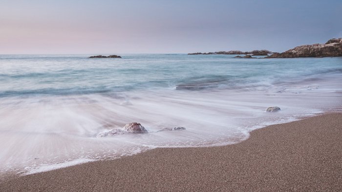 Photo of soft foamy waves on pinkish sand from a crystal blue sea against a pastel sunrise