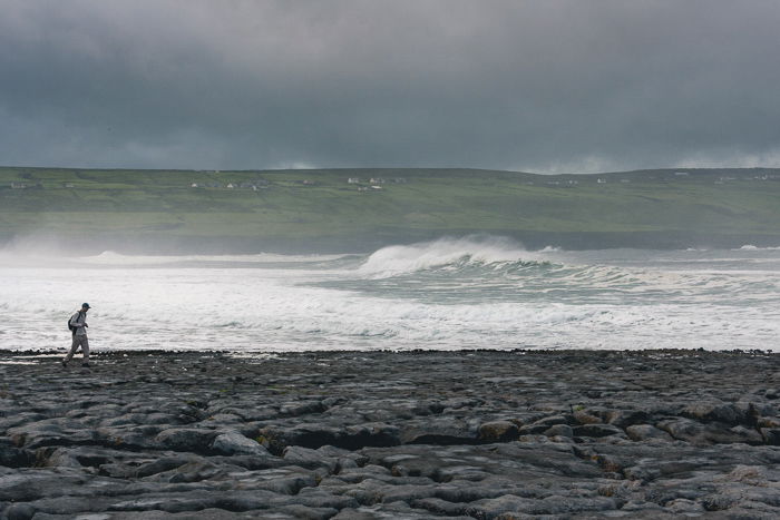 A lone silhouette of a man walking along a beach with big waves behind him.