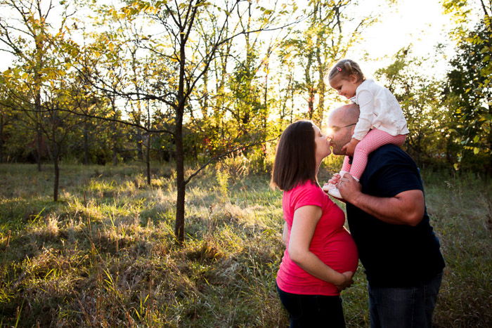 A couple kissing in a forest area, the father holding their little girl on his shoulders