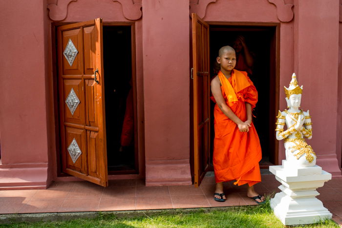 A young novice monk in orange robes standing outside a doorway - figure photography composition 