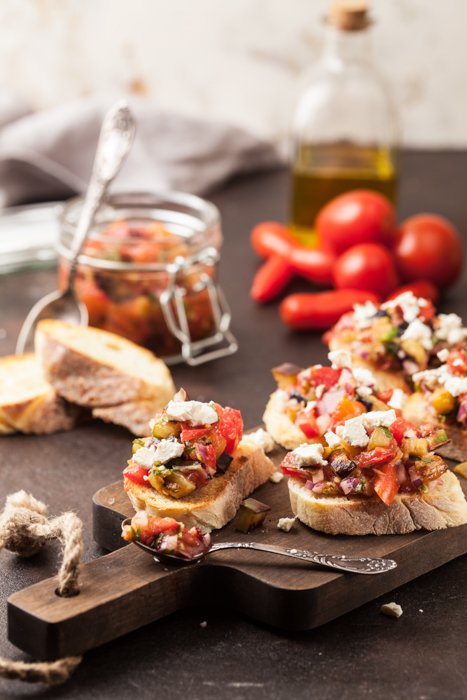 Canapés on a wooden tray, tomatoes, french bread slices, cheese, and a bottle of olive oil on the table.