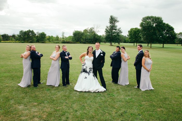 A wedding entourage in a wide grassy field, each couple standing back to back, the bride wearing a white wedding dress with intricate black embroidery