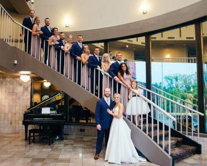 Large group photo of a wedding entourage posing on a big curved staircase, the bride and groom at the bottom in front, a baby grand piano nearby