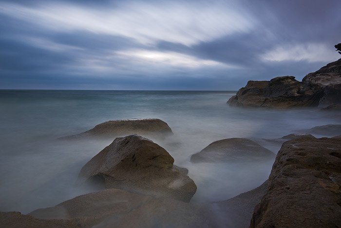 A long exposure ocean photography shot of rocks showing up through a foggy sea, a cloudy sky overhead