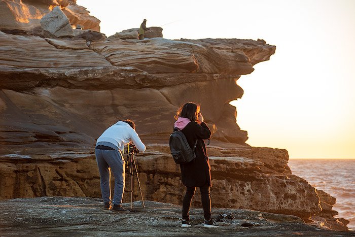 man and woman standing at a rocky cliff, setting up camera and tripod to photograph a seascape