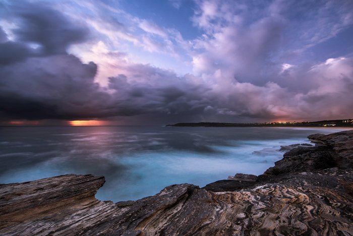 long exposure photo of a seascape at dusk. Blue seas with a rocky shore, reflecting the purple sky and clouds, the orange sunset on the horizon