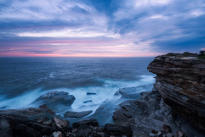 clifftop seascape. rocky cliff over looking a blue sea and foamy waves, a cloudy sky and purple pink sunrise on the horizon
