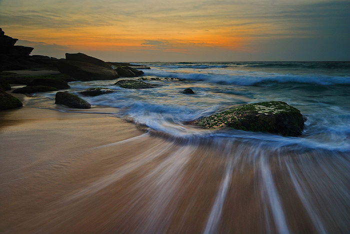 sunset seascape. water trails on the shore, rocks showing up in the foamy waves, golden sunset on the horizon