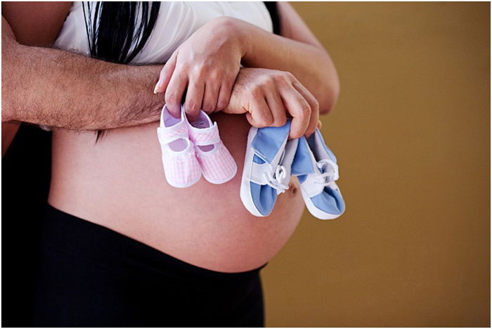 close up couple hugging pregnant belly, holding pink and blue baby shoes