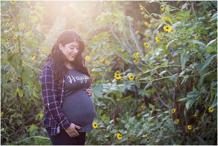 Beautiful maternity photography shot of a pregnant woman wearing a shirt with Happy written on it, cradling her baby standing in a garden with the golden sun glow behind her