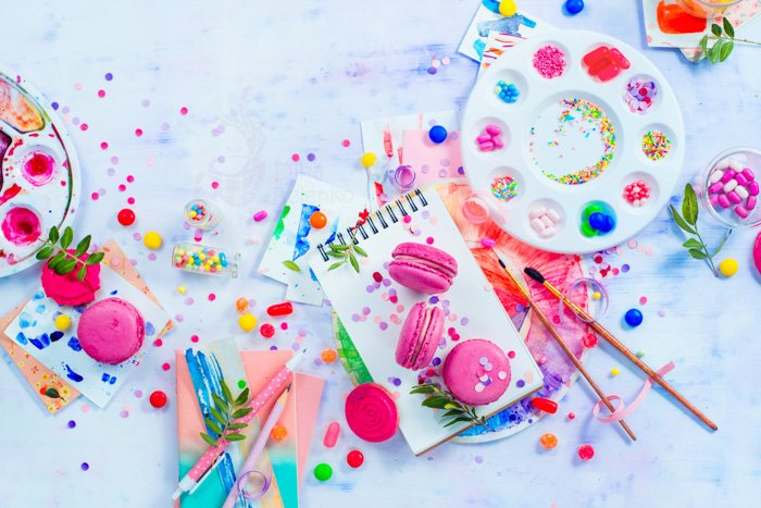 粉红色的马卡龙在艺术家的工作场所五颜六色的平面上放着糖果、糖果和糖屑。用水彩托盘在油漆着色的背景上画一个甜点，里面装满了硬糖果。