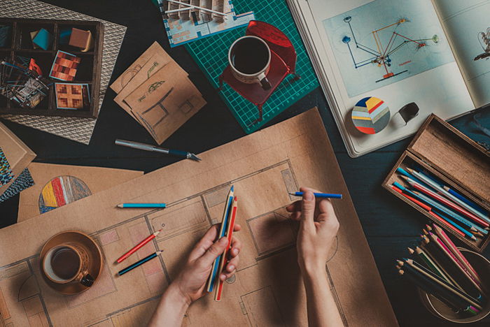 flatlay photo of hands holding drawing pencils, a large brown paper on a table with other drawing materials and coffee