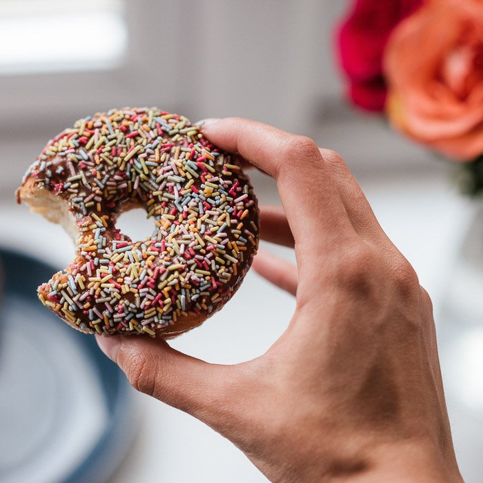 A person holding a chocolate sprinkled doughnut with a bite taken out of it