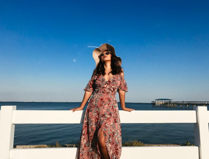 一个女人穿着夏日长裙，戴着太阳镜，戴着大太阳帽，倚在白色的篱笆上，身后是蓝色的大海和天空