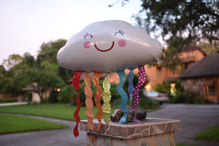 a balloon shaped like a cloud with colourful streamers and a smile, floating in a residential neighbourhood