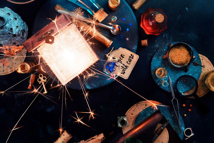 A flatlay of a wooden box with vintage items like paper tag, vials, tools, on a dark grey surface, sparkler lit in the box