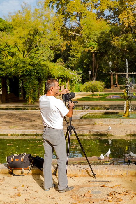photo from behind of a man setting up a DSLR on a tripod by a pond with white birds, the trees in the distance - travel safety tips 
