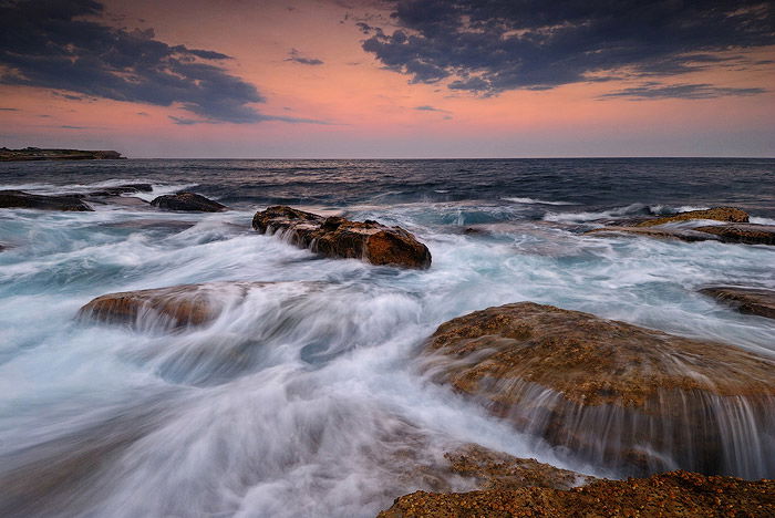 waters rushing over flat brown rocks at Coogee Beach, Sydney, Australia, an orange sunset sky on the horizon