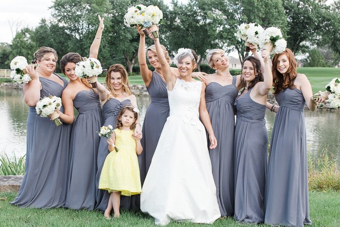 bride with flower girl in yellow and bridesmaids in grey gowns standing in front of a lake smiling and holding their bouquets up