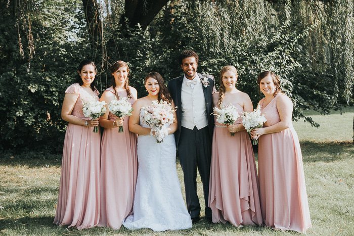 desaturated photo of bride, groom, and bridesmaids in pale pink standing against a backdrop of trees.