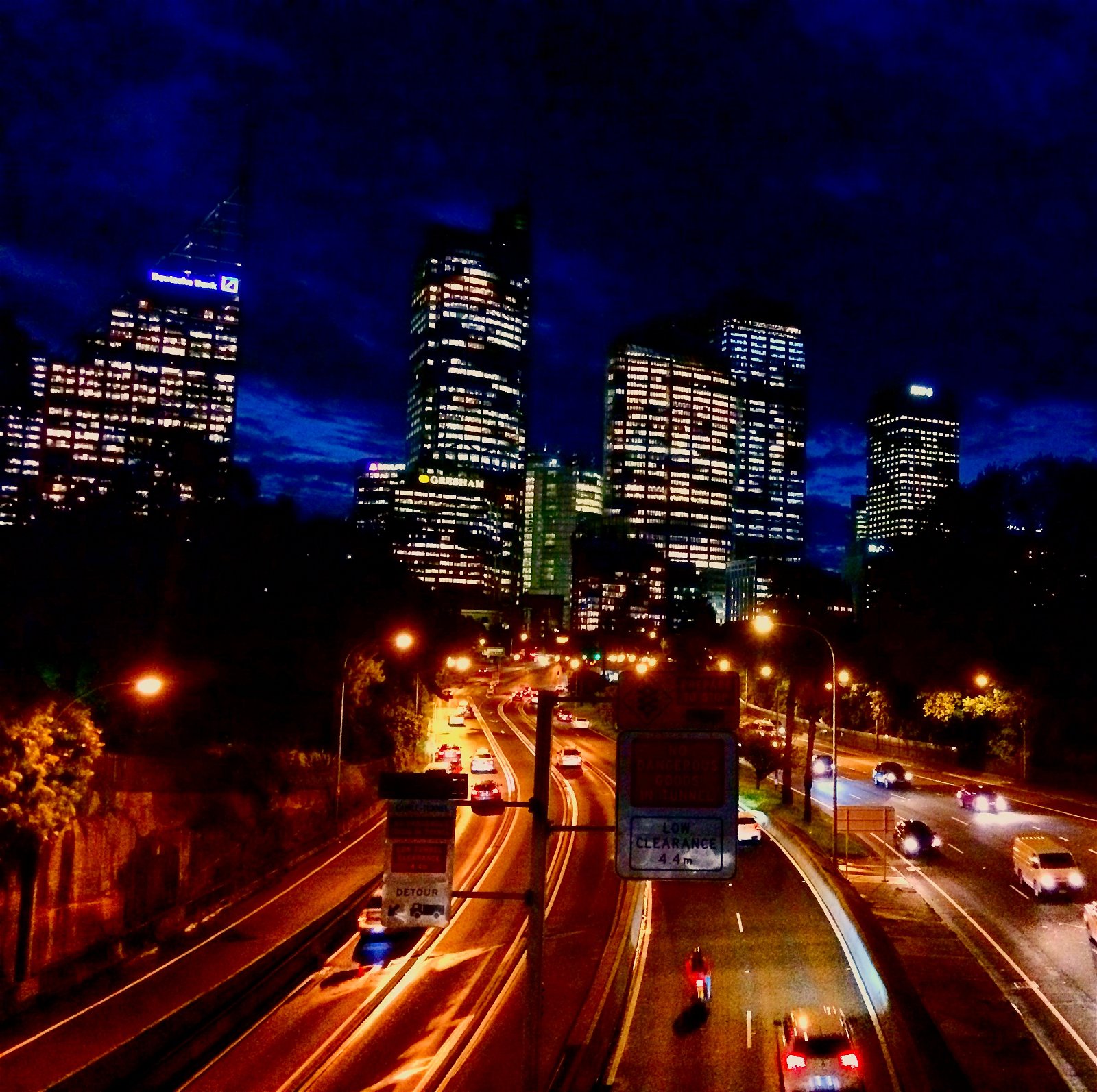 A night scene with three lanes of city traffic and lit streetlight an buildings in the background with a dark blue sky