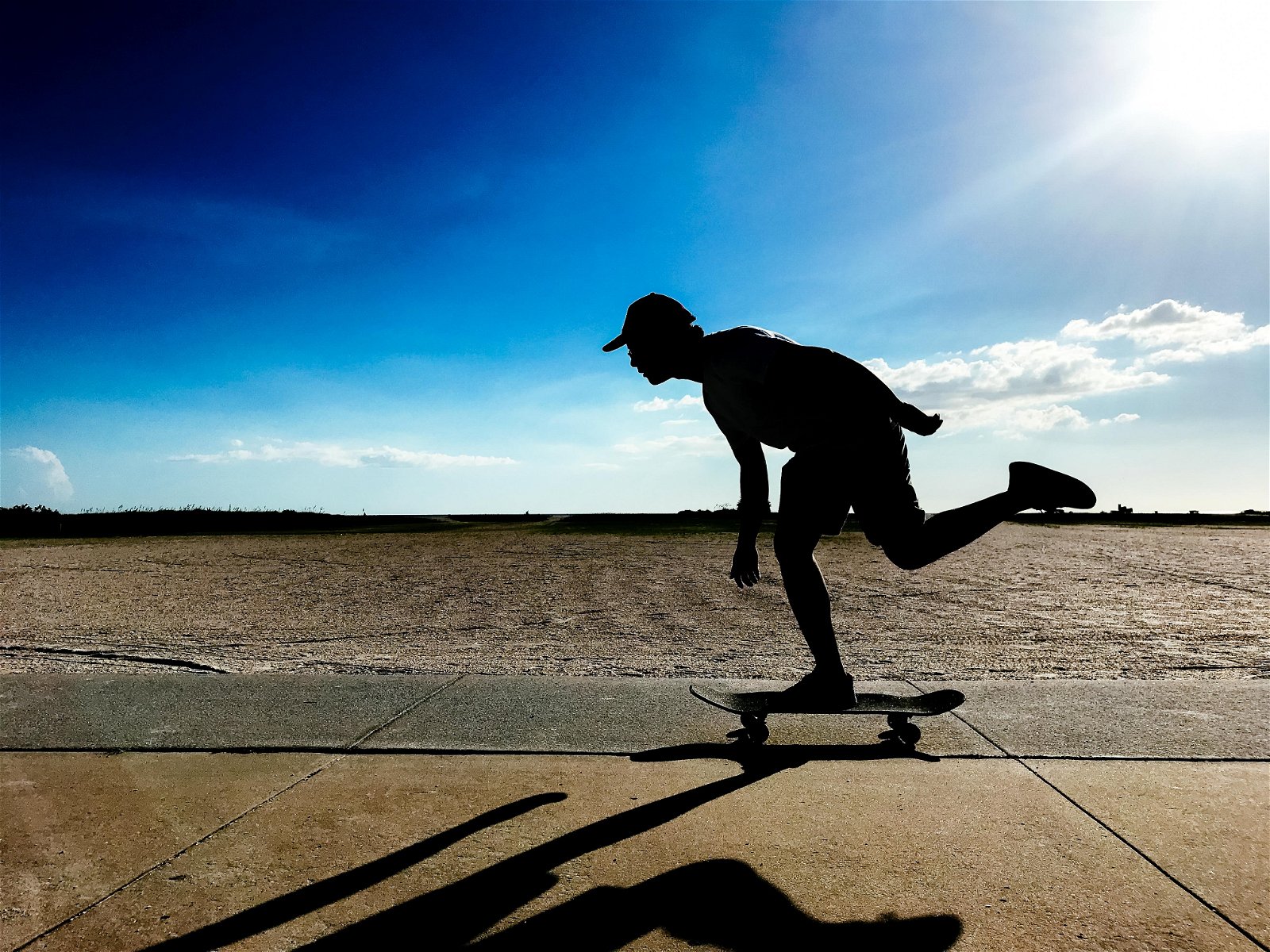 A silhouette and shadow of a skateboarder racing down a sidewalk on a sunny blue-sky day