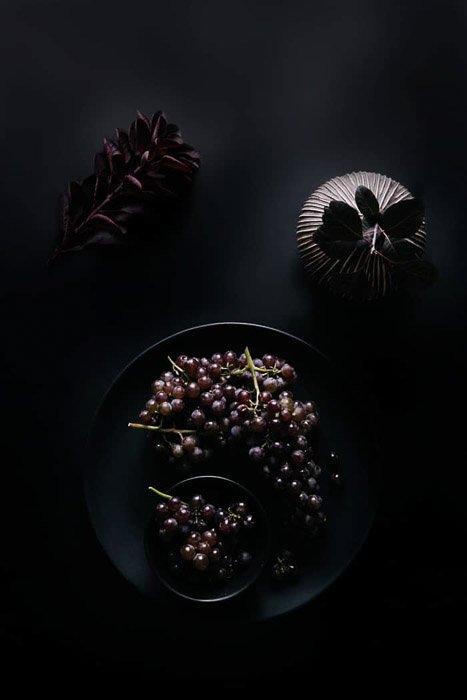 Overhead food shot of a bowl of grapes and other props on a dark background