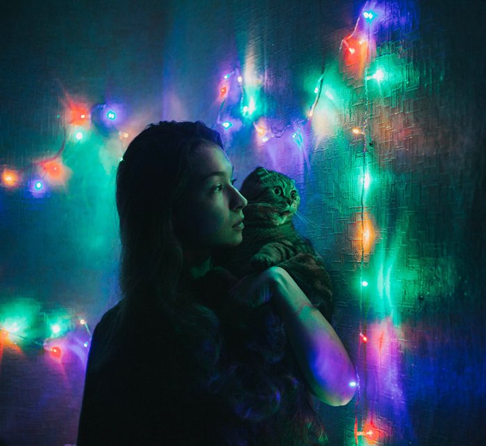 A nighttime portrait of a girl holding a cat surrounded by colorful Christmas lights