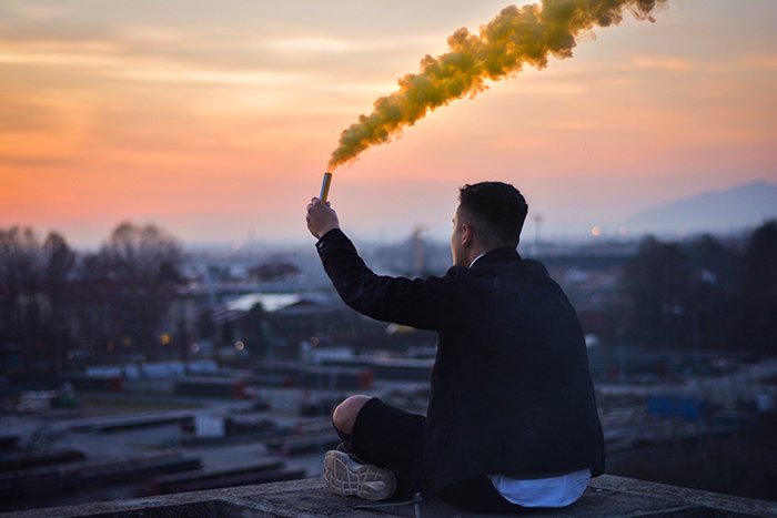 A man sitting on a rooftop holdinhg yellow smoke grenades for photography