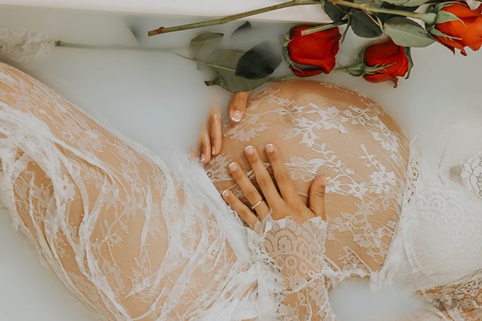 A close up Milk bath photography shoot of a pregnant female model wearing white lace and holding her baby bump, surrounded by flowers