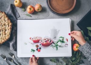 hands holding photo prints of food over a desk with apples and a pie