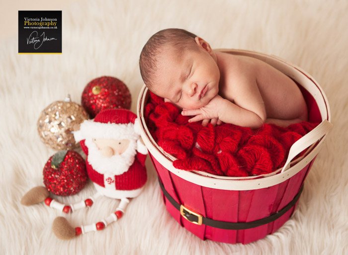 A newborn in a red basket surrounded by Christmas themed newborn photography props