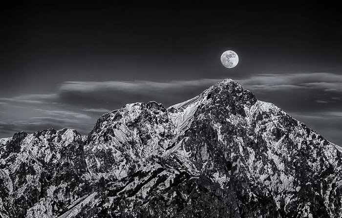 Black and white shot of the full moon over a stunning mountainous landscape 