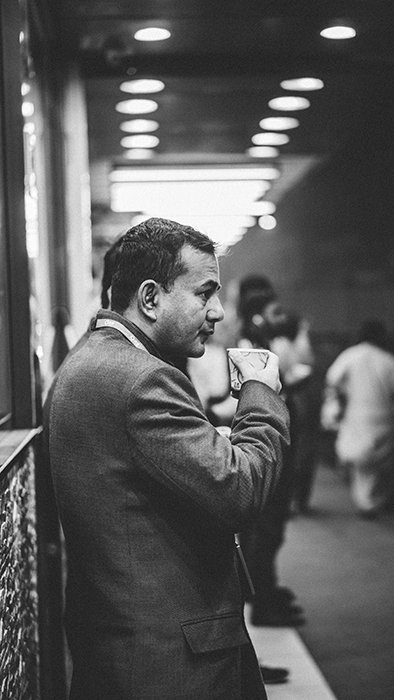 A black and white candid street photography shot of a man drinking coffee at a railway station 