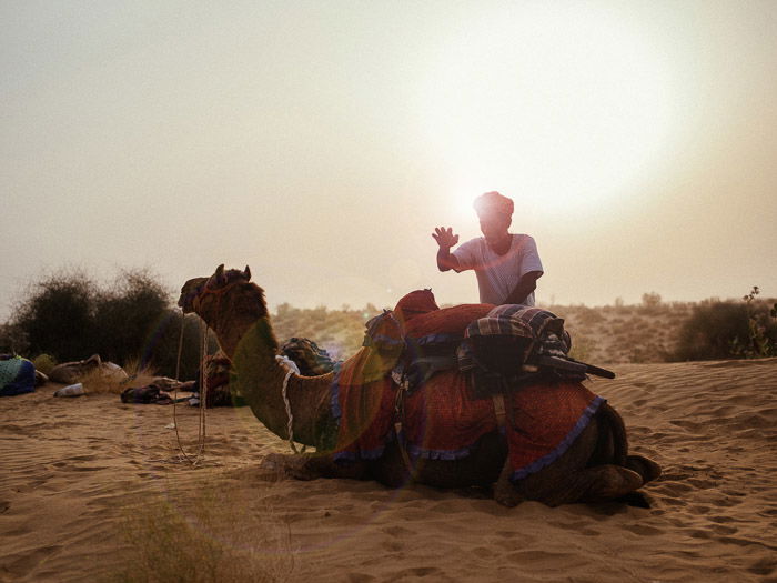 A portrait of a man standing by a camel in the desert, with a beautiful lens flare effect behind him