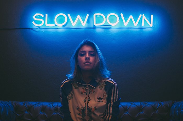 A female model sitting under blue neon signs reading 'slow down'.