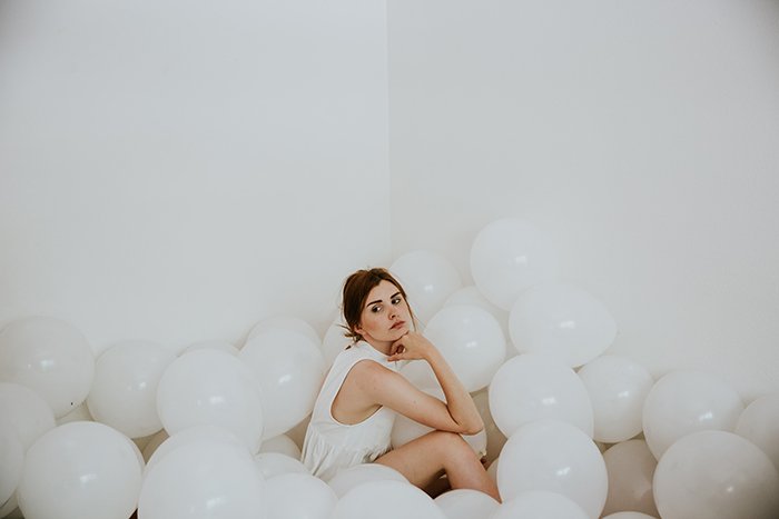 A portrait of a female model sitting in a white walled room, surrounded by white balloons