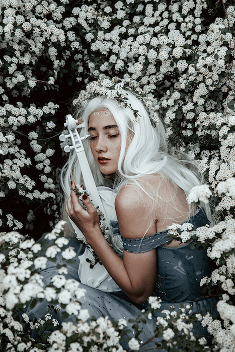 uma imagem de retrato sonhadora de uma mulher de cabelos brancos agachada em um campo de flores brancas segurando um violino branco