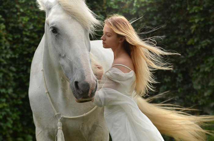 A dreamy photo of a female model with long blond hair posing beside a white horse