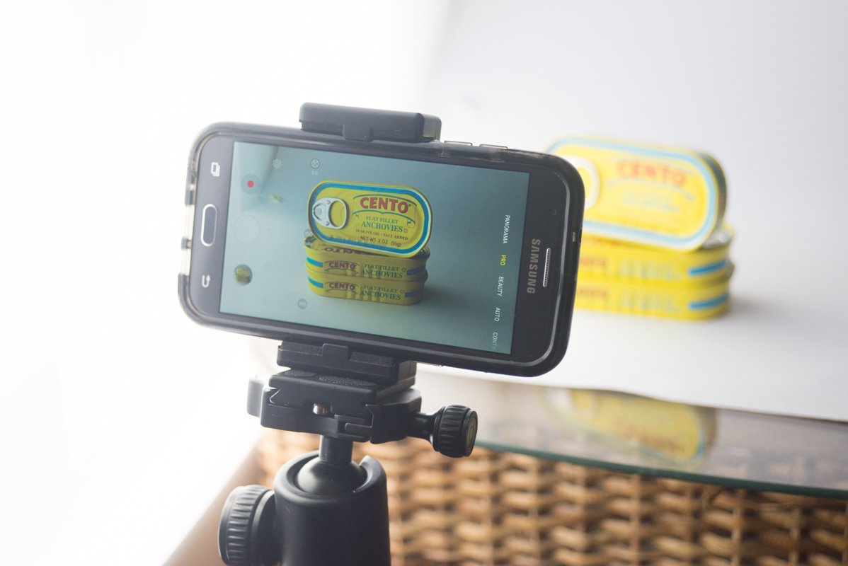 A smartphone set up on a tripod mount to take a product photography shot of cans of food