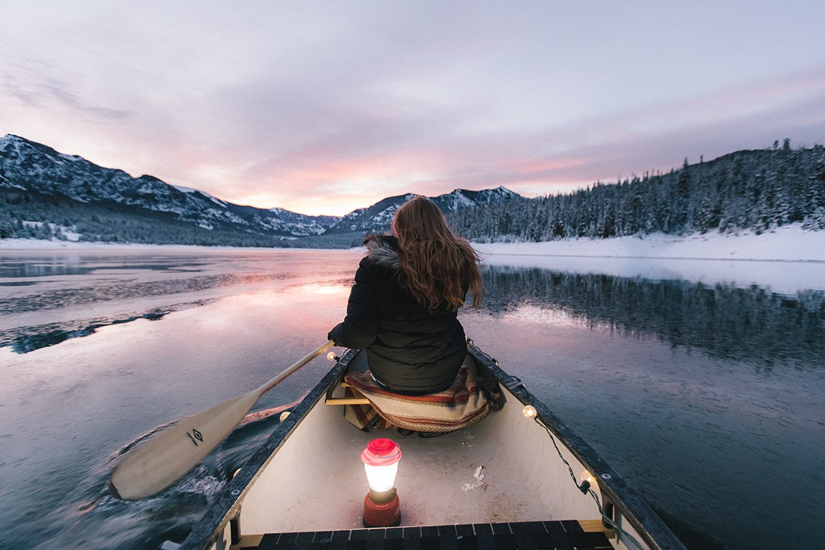 A lifestyle portrait taken from behind a woman in a canoe rowing outside on a lake in winter landscape with an oar