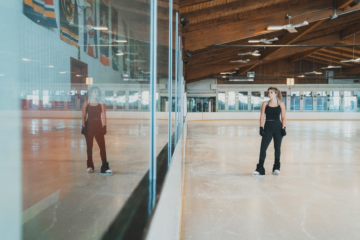 A figure skater in an indoor rink with her reflection in the glass as an example of figure skating photography