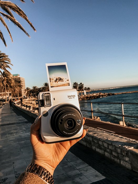 A persons hand holding an instax camera outdoors by the coast