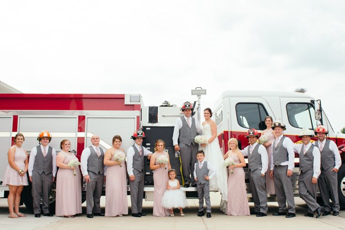 A wedding group photo posed in fireman helmets by a firetruck