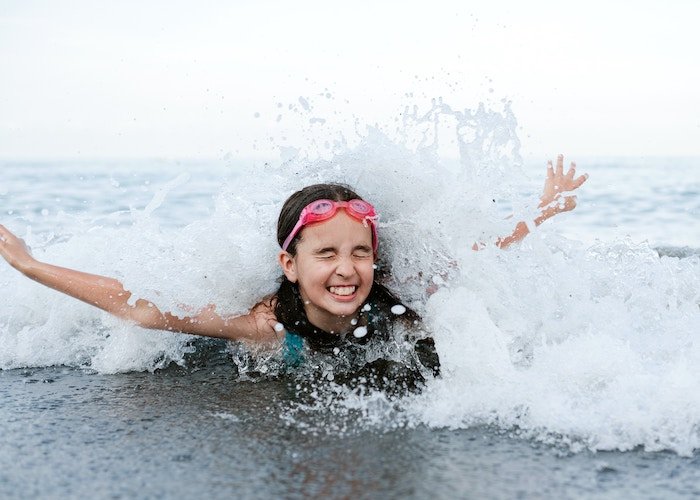 A girl with water googles on her head and her eyes closed being splashed by an ocean wave
